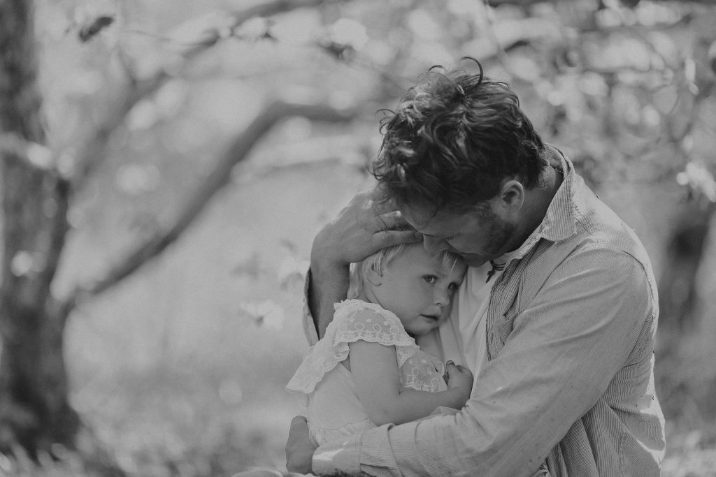 A Good Father is holding and comforting his csad and pouting baby daughter outside on a spring day.